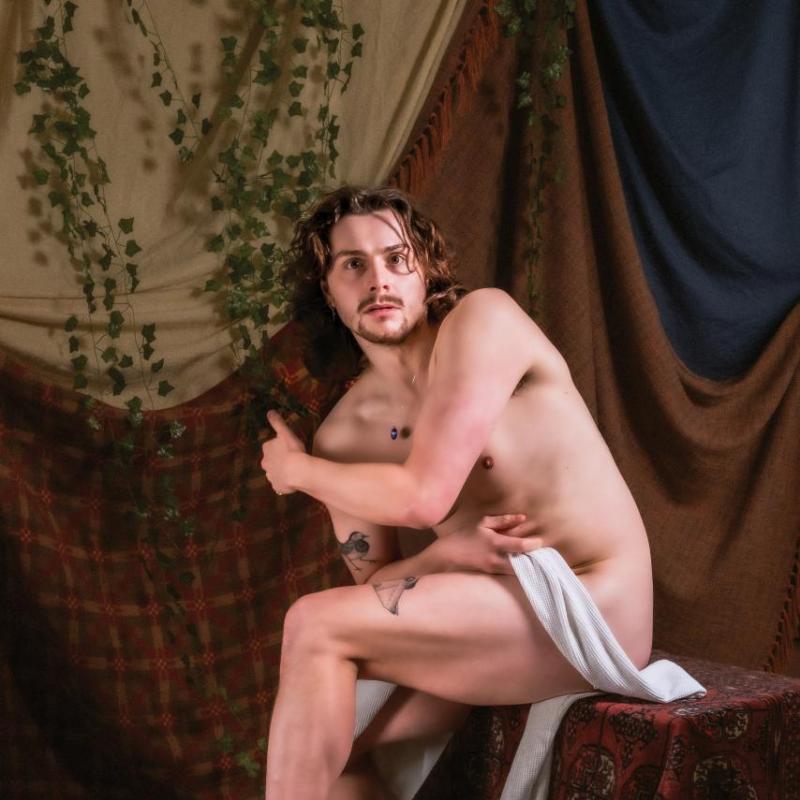 Sam, shirtless, stares into the distance behind the camera. There are vines and curtains draped in the background.