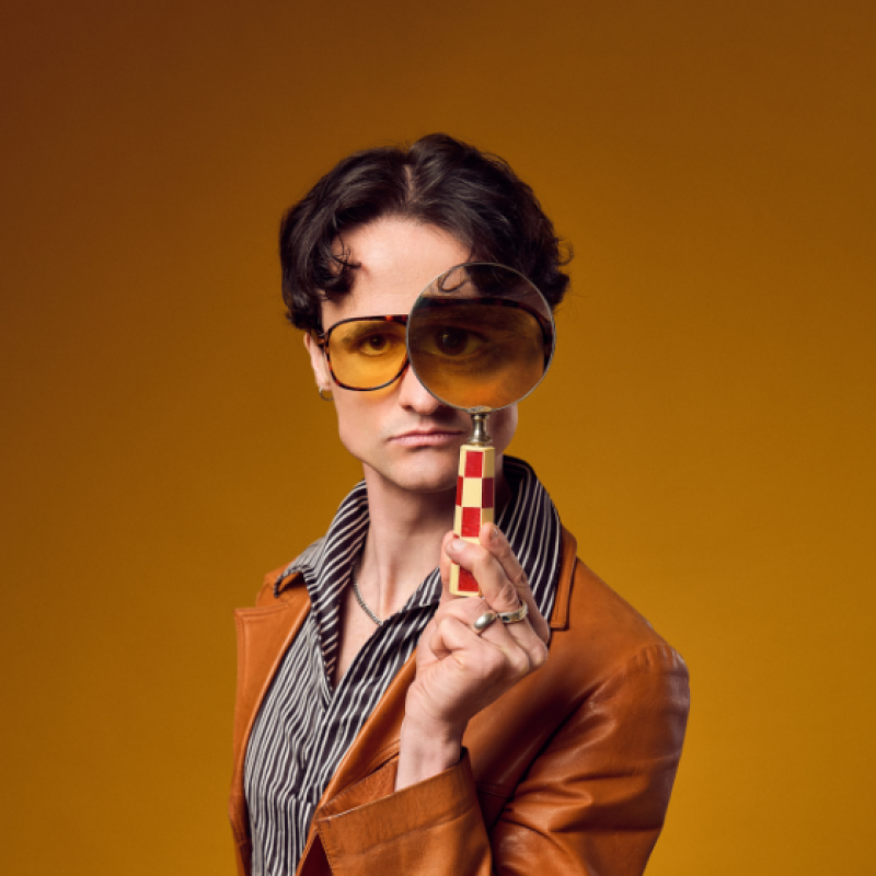 Jazz holding a magnifying glass up to his eyes wearing a 70s inspired outfit