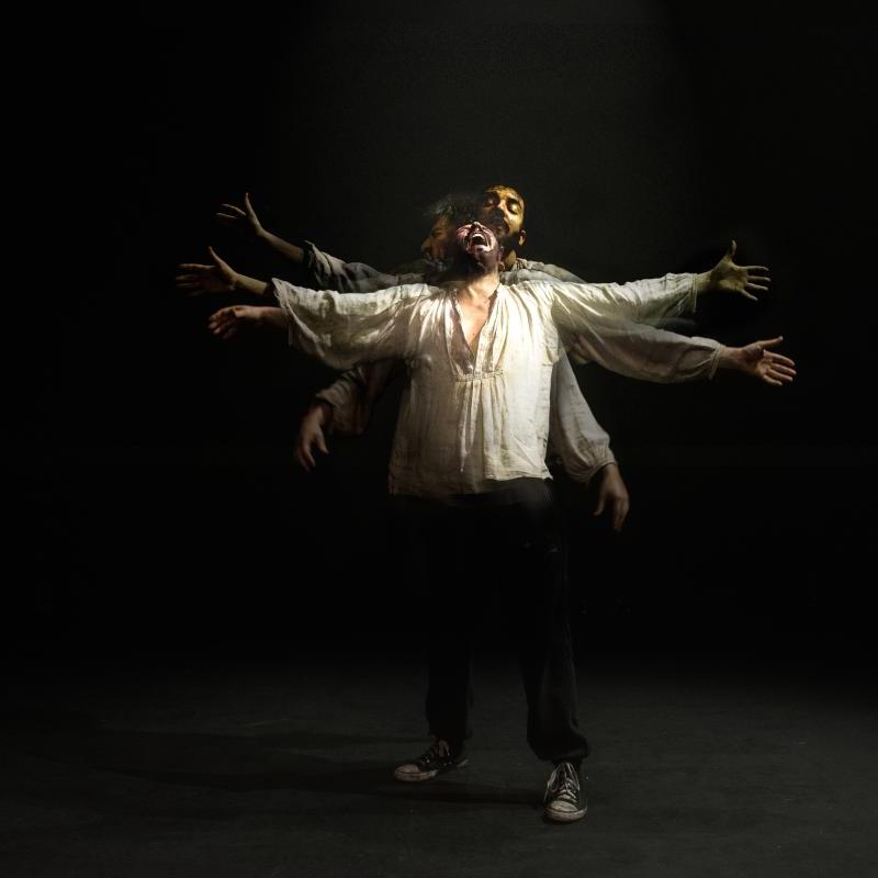 A performer in a loose white shirt stands with their arms outstretched, against a black background on an empty stage. They appear to be crying to the heavens. There are faint images of them in very slightly different poses superimposed on this main one. The effect means that it looks like they have six arms, and three heads, all doing something very slightly different.