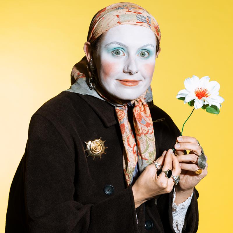 A person with white make-up wearing a headscarf, rings and holding a flower looks out at the viewer.