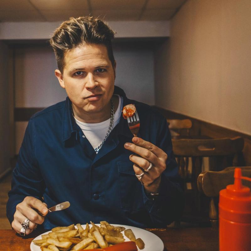 Luke Wright sits at a table of what looks like a greasy spoon type restaurant. They're holding up a fork that's got sausage and ketchup on it, and smiling at the camera with their mouth closed, mid-chew.