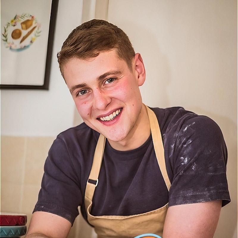 A smiling Peter covered in flour smiles at the camera holding a whisk.