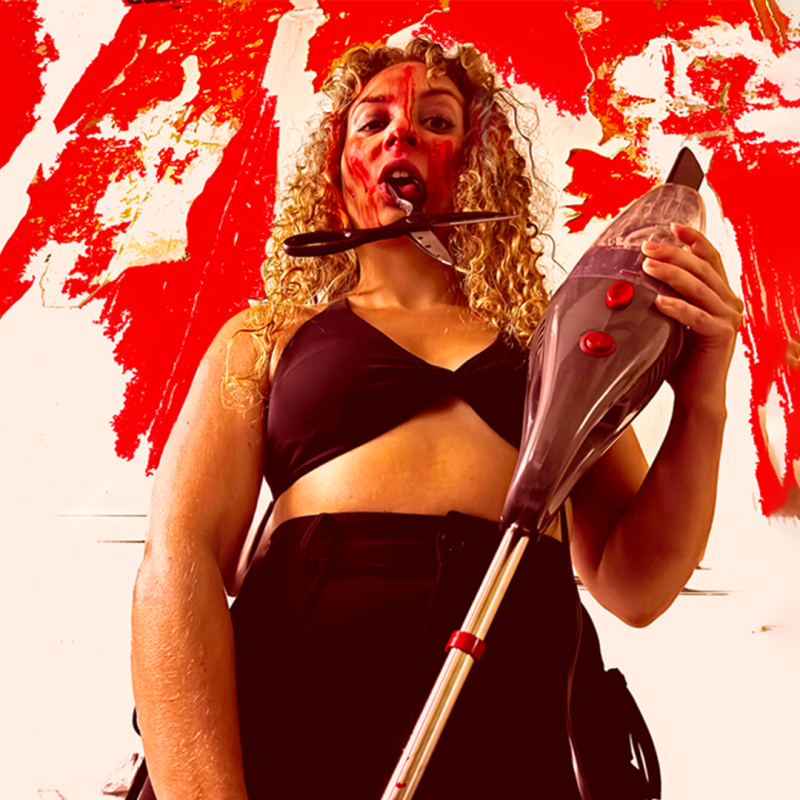 Performer with scissors hanging out of their mouth, holding a vacuum clear with blood over their face.