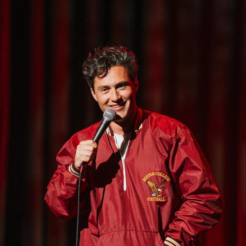 Alec wearing a red varsity style jacket holding a microphone on stage