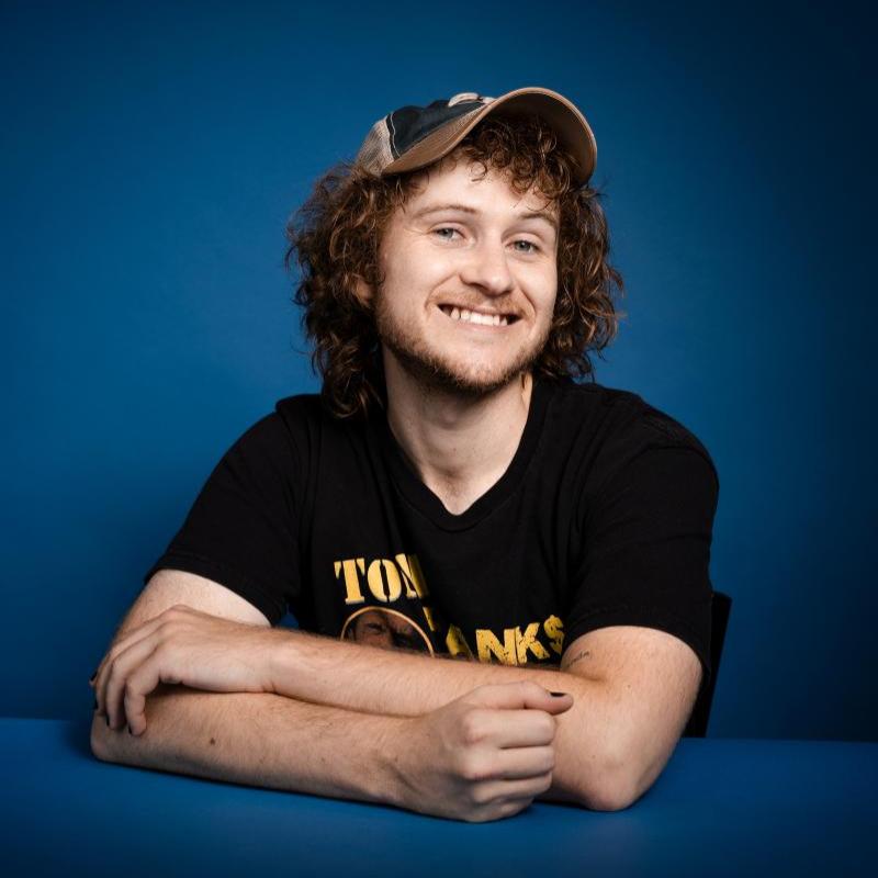 Casey smiles with arms loosely folded wearing a cap.