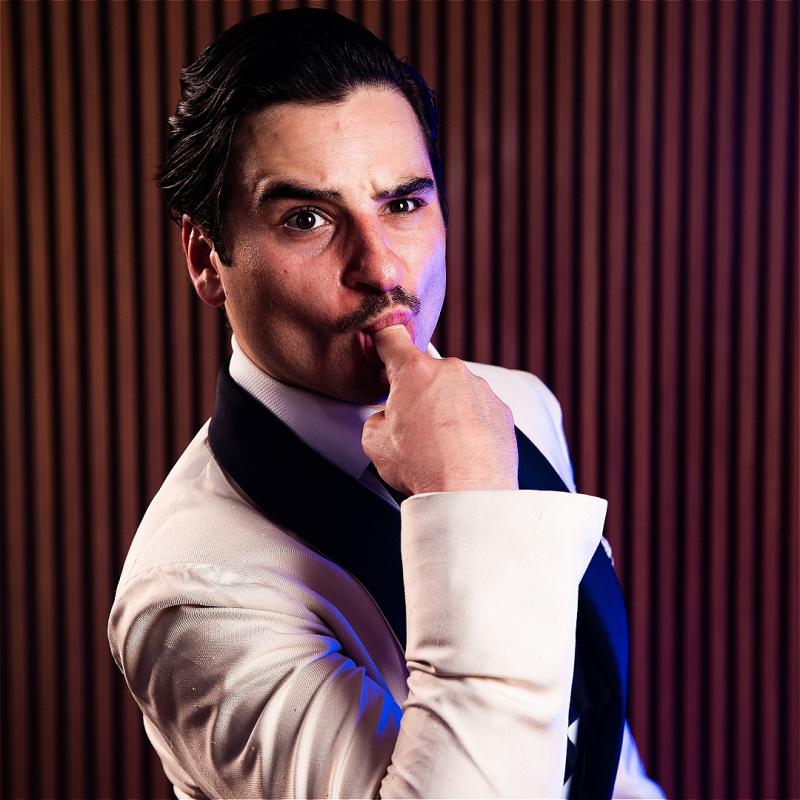 Joey wearing a white tuxedo sticks a finger in his mouth and raises an eyebrow
