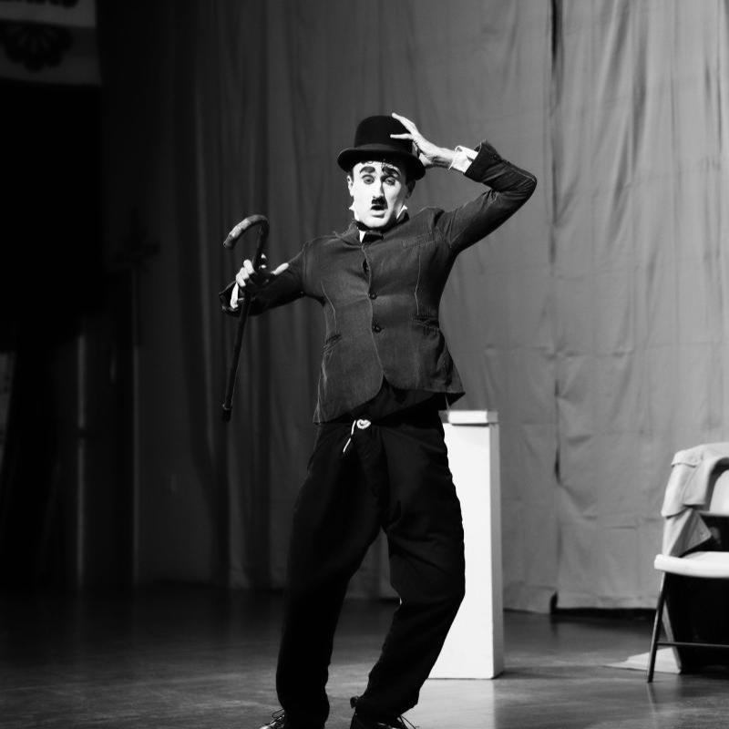 A black and white full body shot of a man dressed as Charlie Chaplin. He has a white face, black eyebrows, eyeliner and toothbrush moustache, and wears a tight black jacket, baggy pants and bowler hat and holds a bamboo cane in one hand. His other hand is on his hat. He appears to be trudging into the wind.