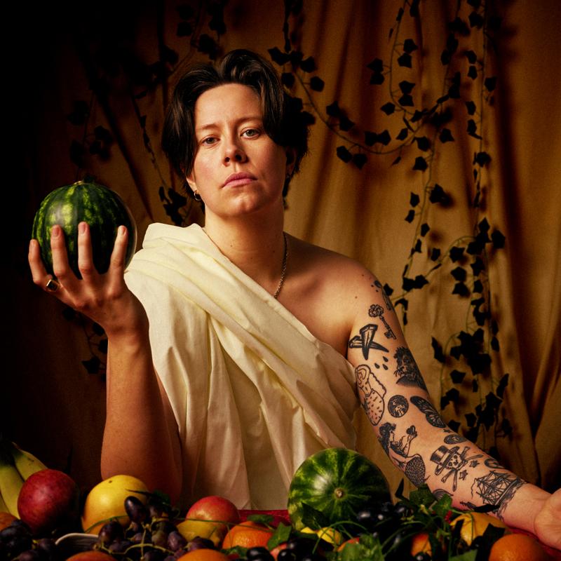 Chloe Petts wearing a toga amidst a lavish table of food and holding a watermelon in one hand