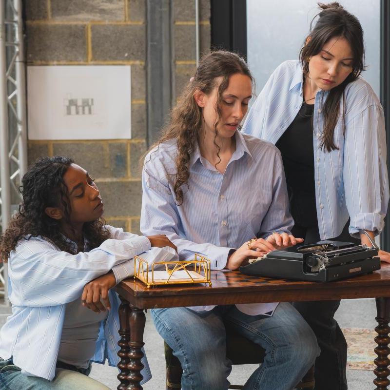 three women sat at a table looking at a typewriter