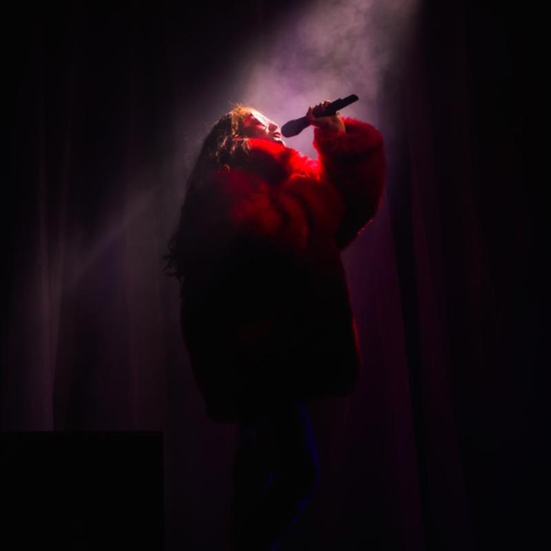 A woman in a red fluffy coat and pink sunglasses poses in a spotlight with a microphone about to sing