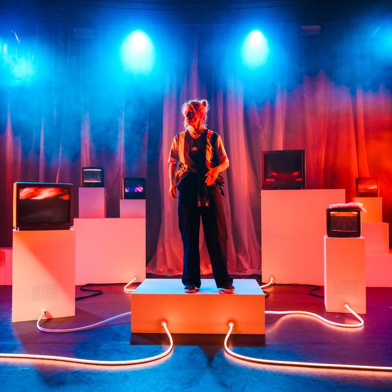 A woman in a football kit stands on a platform. She is surrounded by 90s TVs on plinths