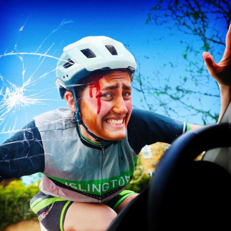 The performer dressed in a cycling outfit with a white helmet on top of a broken car windshield with blood running down their face