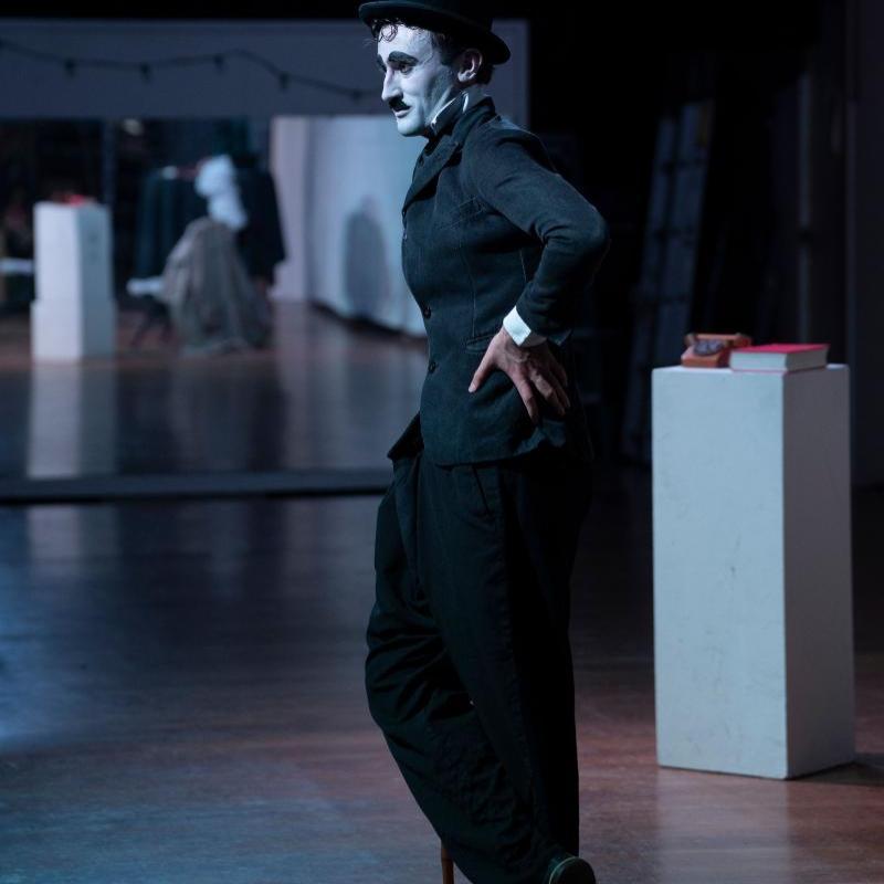 A full body shot of a man dressed as Charlie Chaplin. He has a white face, black eyebrows, eyeliner and toothbrush moustache, and wears a tight black jacket, baggy pants and bowler hat and is leaning with one hand on his cane. His other hand is on his waist and one leg is crossed over the other.