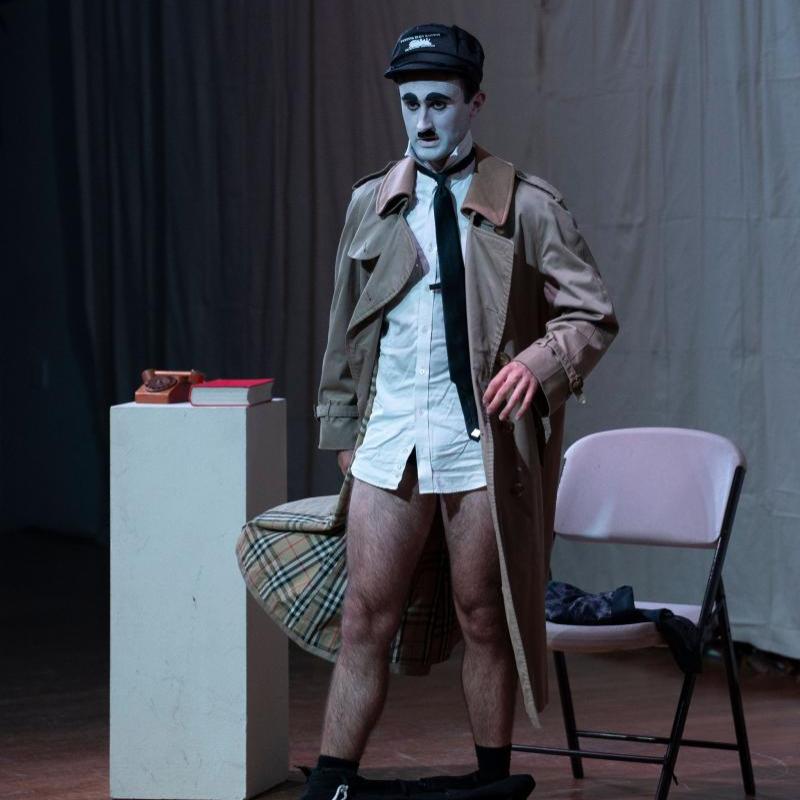 A full body shot of a man dressed as Charlie Chaplin dressed as Hitler referencing Chaplin's 1940 film 'The Great Dictator'. He has a white face, black eyebrows, eyeliner and toothbrush moustache, and wears a white shirt, black tie, cream coloured trench coat and a black military looking cap. His trousers are around his ankles.