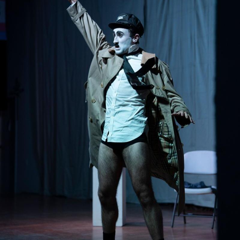 A full body shot of a man dressed as Charlie Chaplin dressed as Hitler referencing Chaplin's 1940 film 'The Great Dictator'. He has a white face, black eyebrows, eyeliner and toothbrush moustache, and wears a white shirt, black tie, cream coloured trench coat and a black military looking cap. His trousers are around his ankles. He is giving a vigorous Nazi style salute.