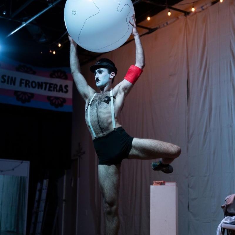 A full body shot of a man dressed as Charlie Chaplin dressed as Hitler referencing Chaplin's 1940 film 'The Great Dictator'. He has a white face, black eyebrows, eyeliner and toothbrush moustache, and wears tight black shorts, white suspenders, black swastika nipple covers, a red arm band and a black military cap. He holds a giant balloon globe above his head and has one leg raised in a balletic position.