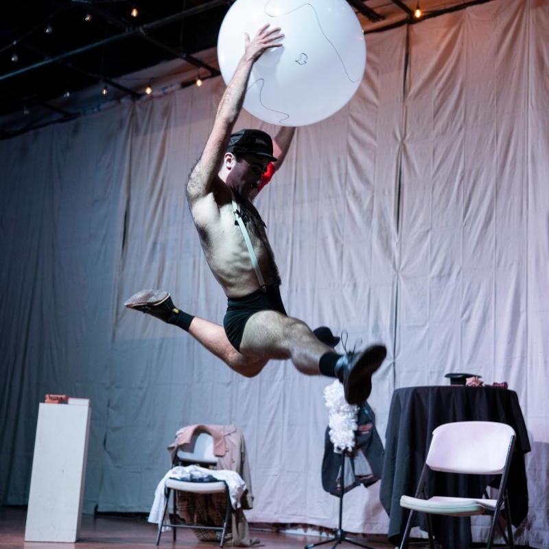 A full body shot of a man dressed as Charlie Chaplin dressed as Hitler referencing Chaplin's 1940 film 'The Great Dictator'. He has a white face, black eyebrows, eyeliner and toothbrush moustache, and wears tight black shorts, white suspenders, black swastika nipple covers, a red arm band and a black military cap. He holds a giant balloon globe above his head and is caught in mid air doing a split jump across the stage.