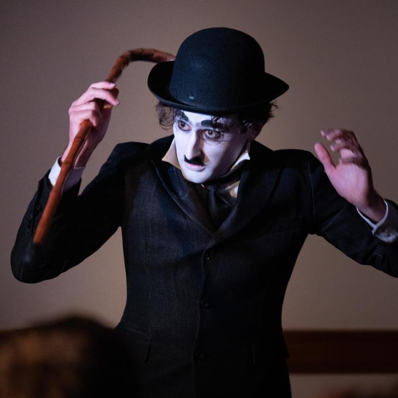 A medium close up of a young man dressed as Charlie Chaplin. He has a white face, black eyebrows and toothbrush moustache, and wears a white shirt, black jacket and bowler hat and holds a bamboo cane in one hand. His arms are raised