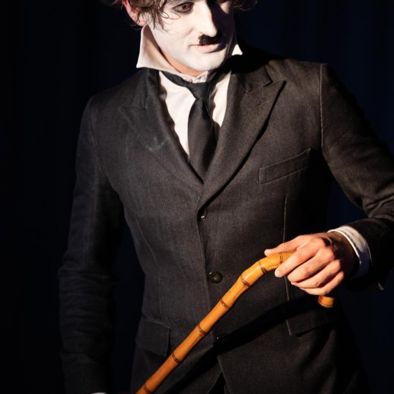 A medium close up of a young man dressed as Charlie Chaplin. He has a white face, black eyebrows, eyeliner and toothbrush moustache, and wears a white shirt, black jacket, tie and bowler hat and holds a bamboo cane in both hands.