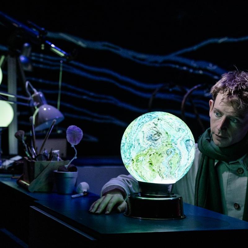 The Little prince - a young boy with blond hair sat on a stool at a desk in his workshop looking at a globe of the world
