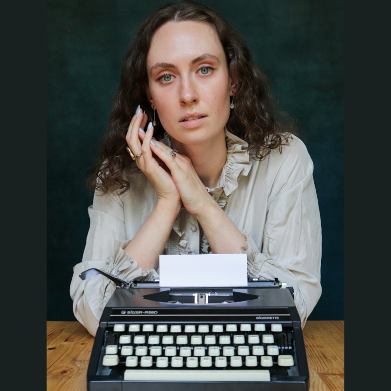 Woman sits behind a typewriter on a wooden desk, dark green background