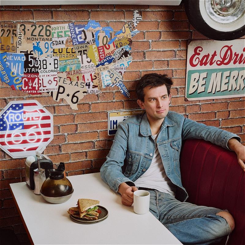 A man in a cafe, holding a mug. There is a sandwich on the table in front of him. 
