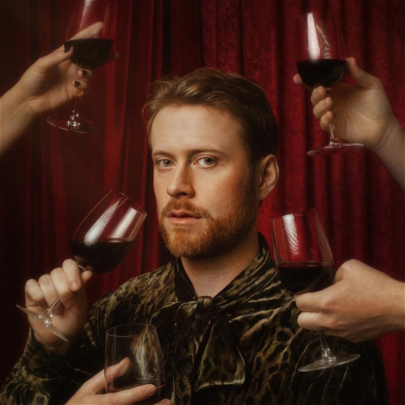 A sultry looking John is surrounded by full glasses of red wine held by outstretched hands.