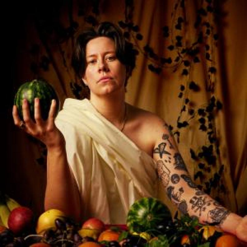 Chloe Petts wearing a toga amidst a lavish table of food and holding a watermelon in one hand