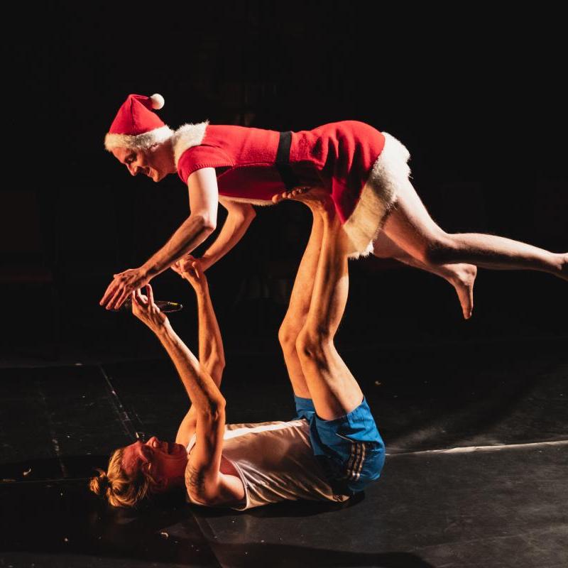 Two performers holding a balancing lift pose, one wears a red and white santa costume.
