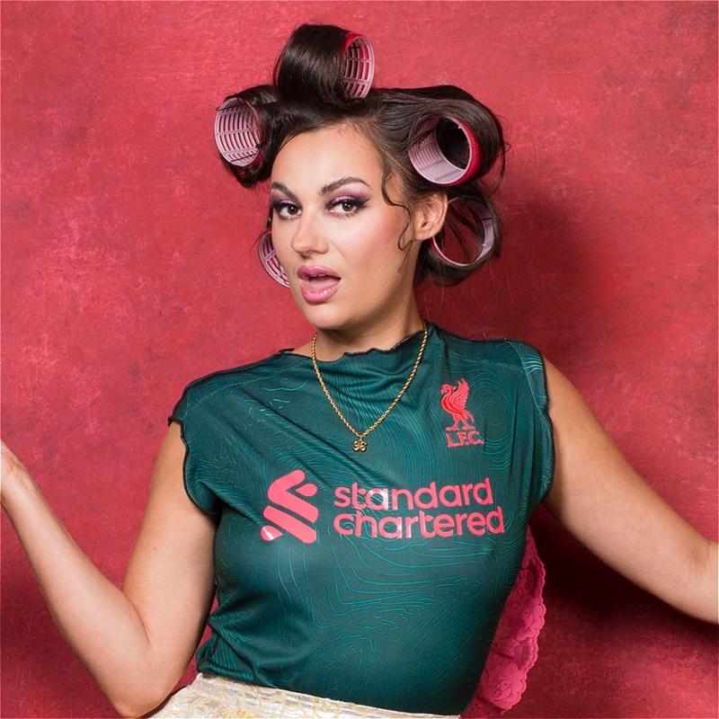 A woman in a Liverpool FC jersey poses playfully with pink hair rollers, against a vibrant red background.