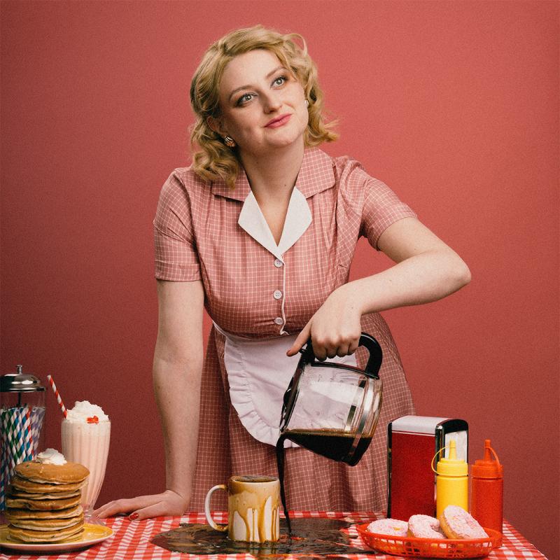 Lorna wearing a red gingham dress with an apron smiling sweetly as she pours coffee into an overflowing cup.