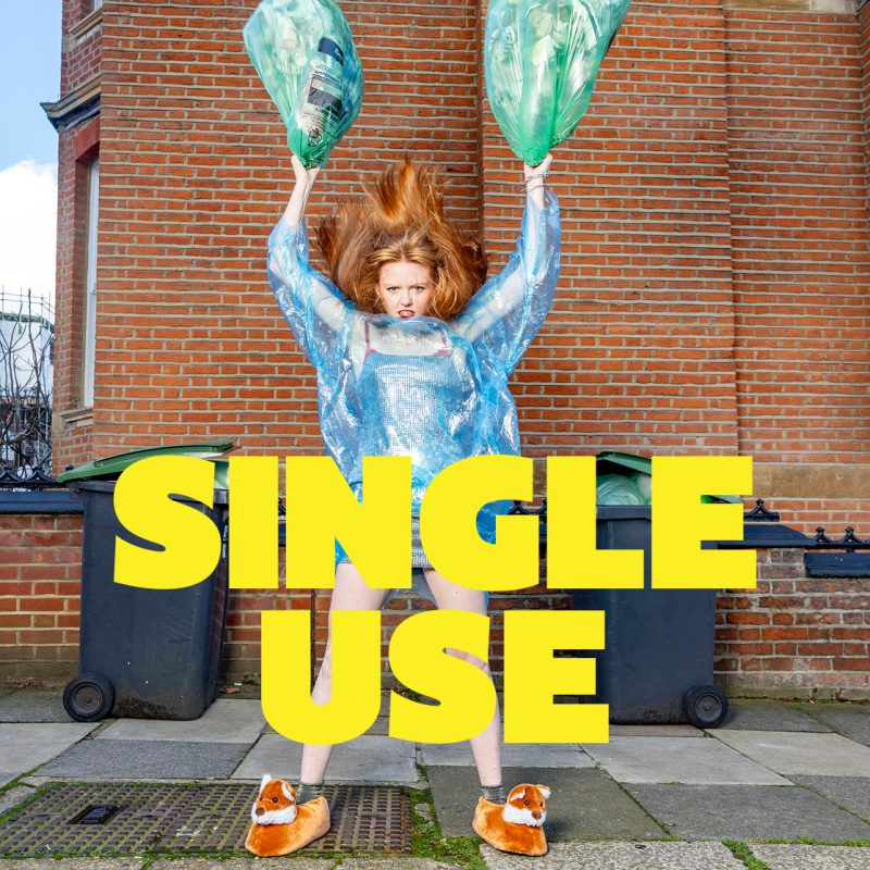 A red haired lady looks confused at the camera with a big blue recycling bag draped across her body. In her hands she hoists two full green recycling bags above her head in a menacing position.A red haired lady looks confused at the camera with a big blue recycling bag draped across her body. In her hands she hoists two full green recycling bags above her head in a menacing position.