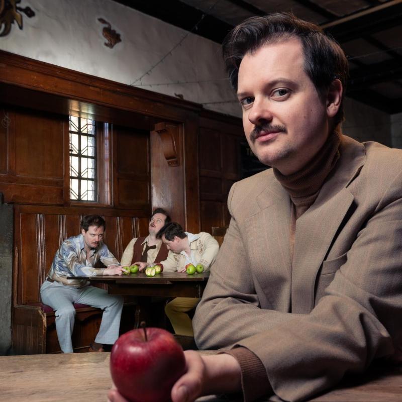 Andy in a pub wearing brown suit holding an apple at the forefront. In the background, Andy is three different drunk people.