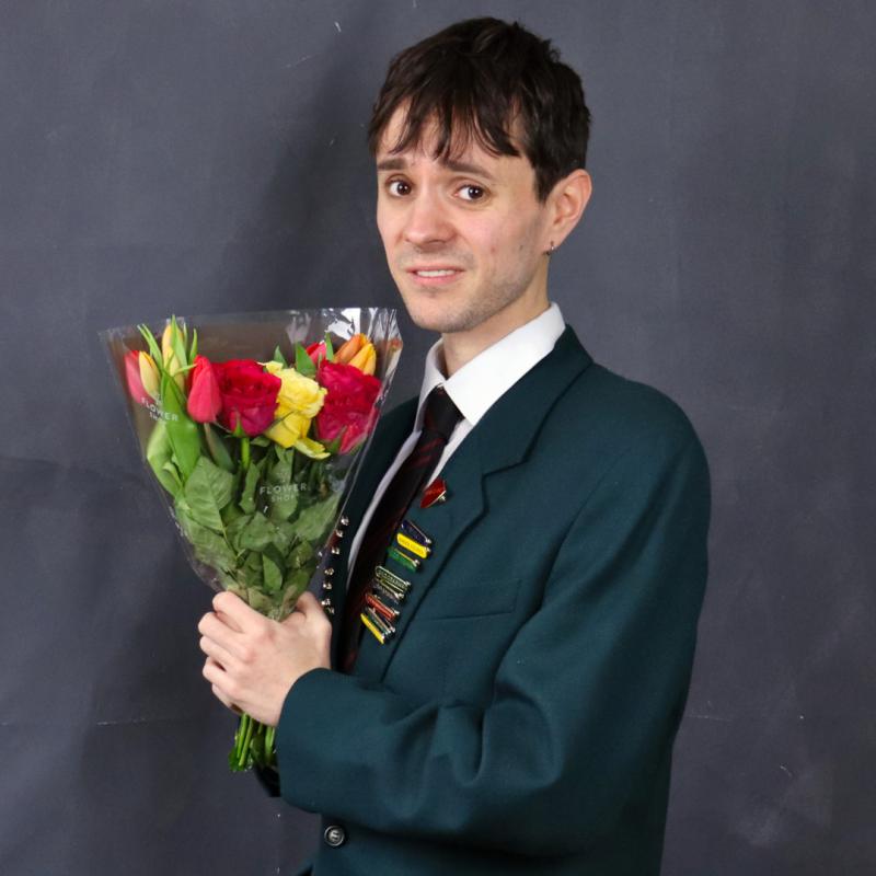 A boy in a school uniform blazer, and lots of achievement pins, holds a bunch of flowers and looks apologetic