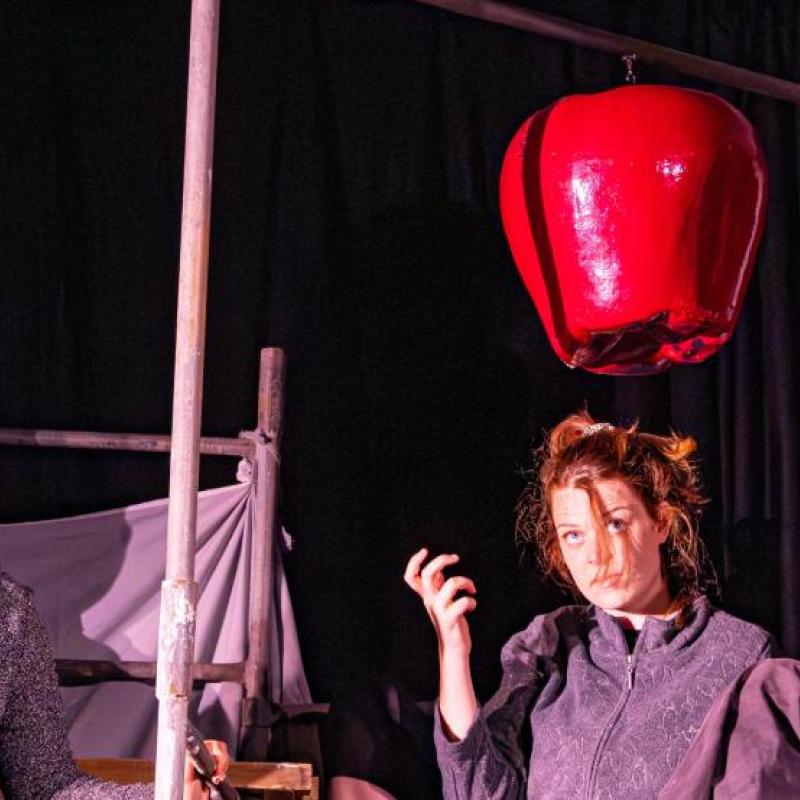 4 actors holding a metaphorical apple wearing grey jumpsuits whilst a large prop apple dangles above them.