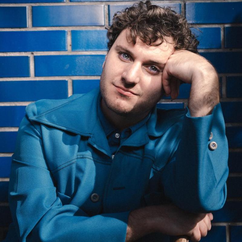 Headshot of performer wearing all blue set against a tiled blue background.