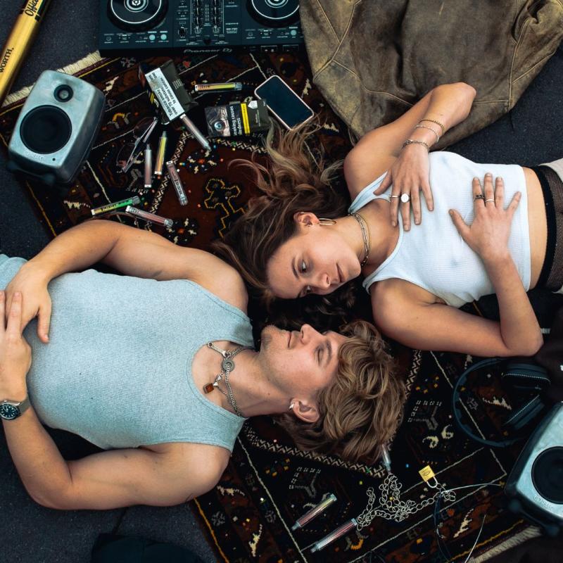 A couple lay on a rug, wearing vests and looking at each other. There is a DJ Deck, Speakers, and other techno items around them