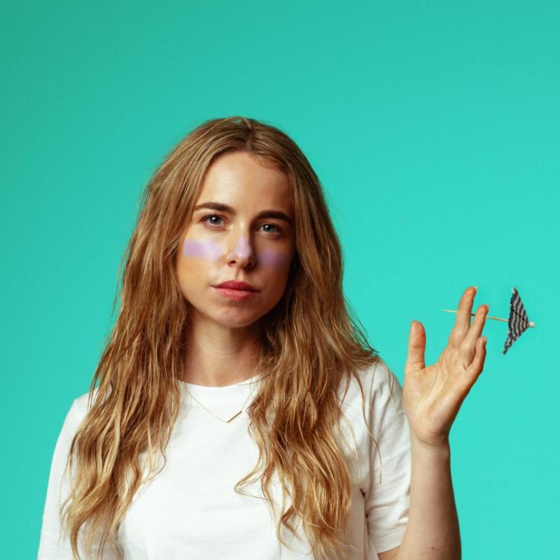 Maisie stands on a light blue background in a white tshirt. With purple warpaint on her face, and a cocktail umbrella in her hand. 