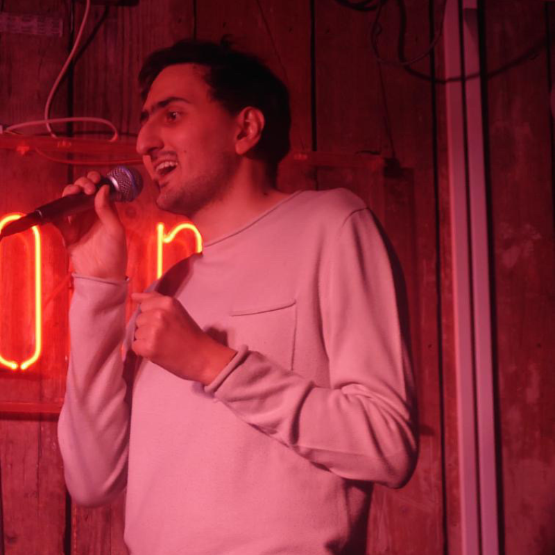 Aarian holding a mic and doing stand-up in pink lighting