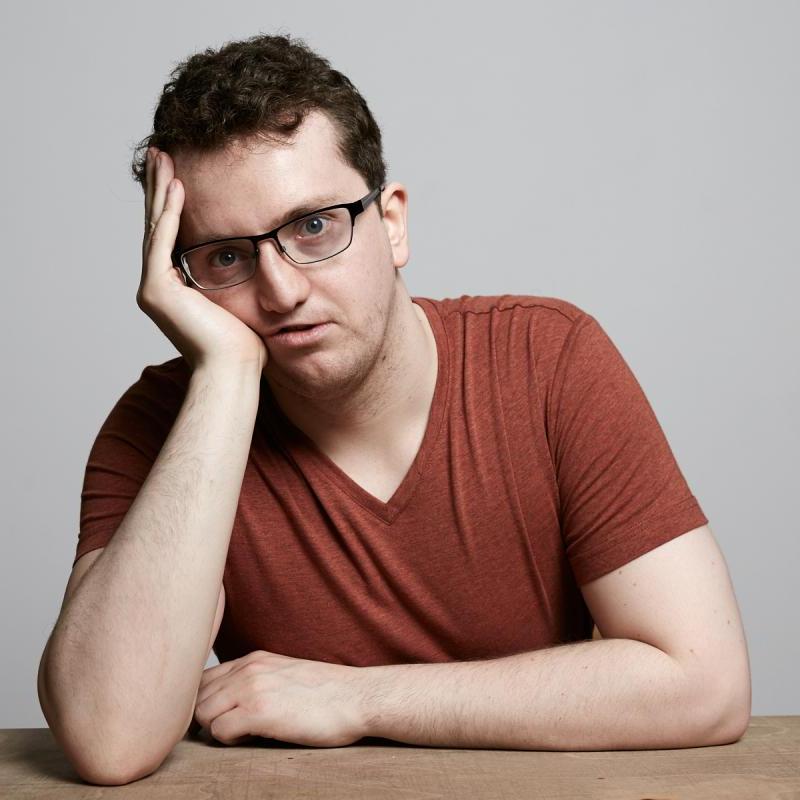 Jonny resting his face on his hands on a wooden table and a grey background.