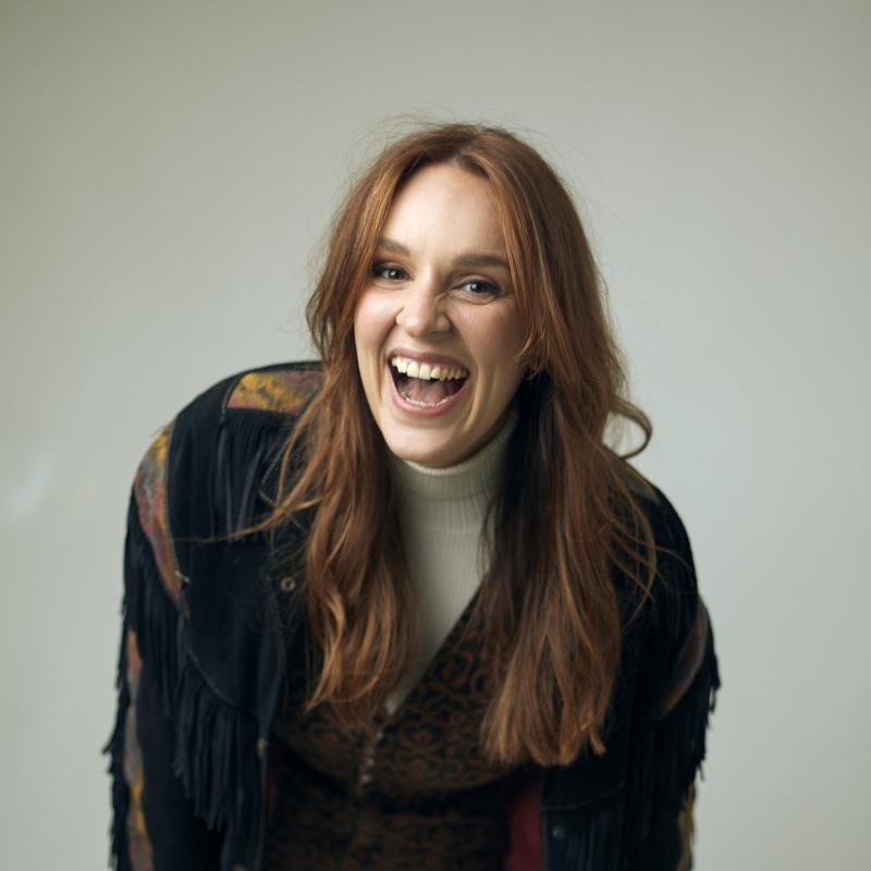 Katie Norris wearing a fringe jacket leans forward and laughs to camera
