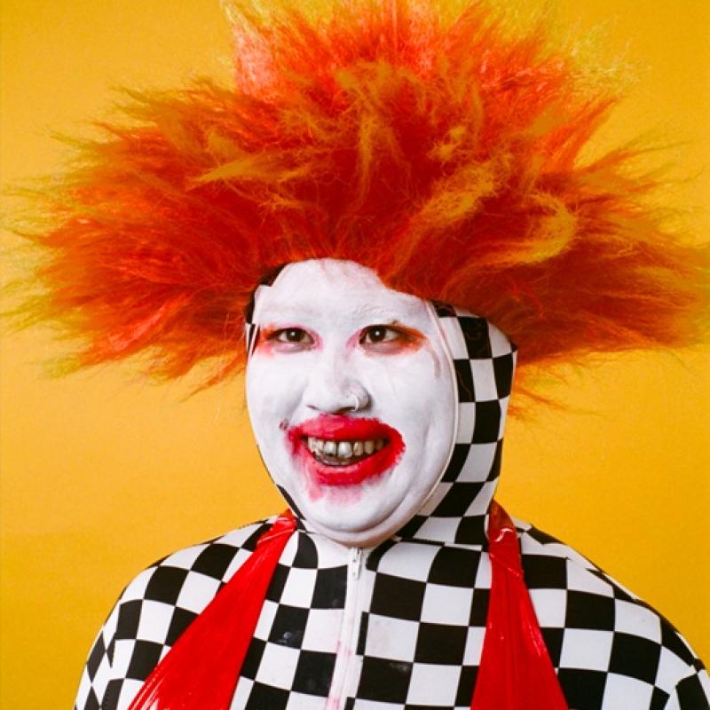 Su Mi with white face paint and red lipstick with a wild orange spiky wig. Wearing a full checkerboard one-piece with a red latex swimsuit.