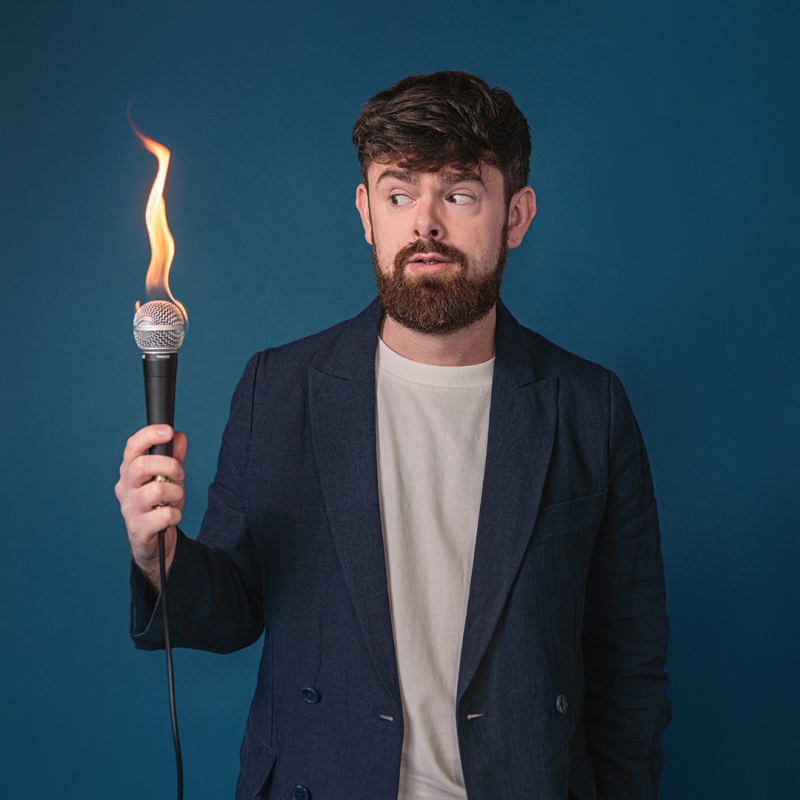 Tom, wearing a blue suit and white tshirt, holds a flaming mic in his right hand and is looking at it concerned. 