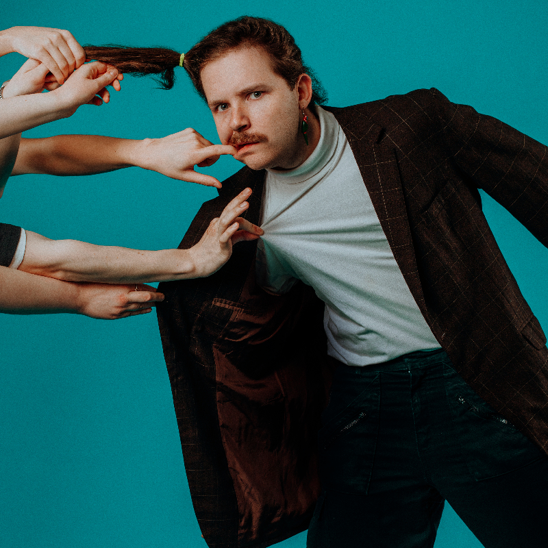 Performer in a suit is having their hair in a pony tail pulled by five different hands.