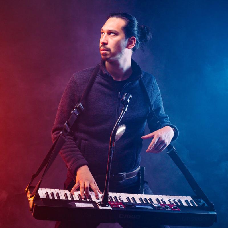 Huge Davies, wearing a keyboard, looks fiercely to the side framed in pink and blue light