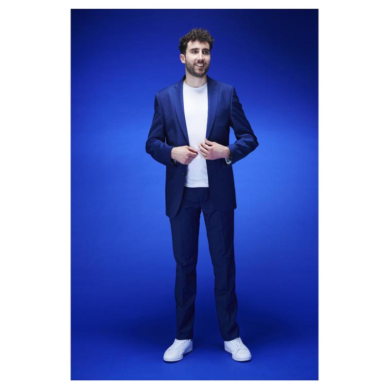 Steve stands in a blue suit, and white t-shirt on a blue background