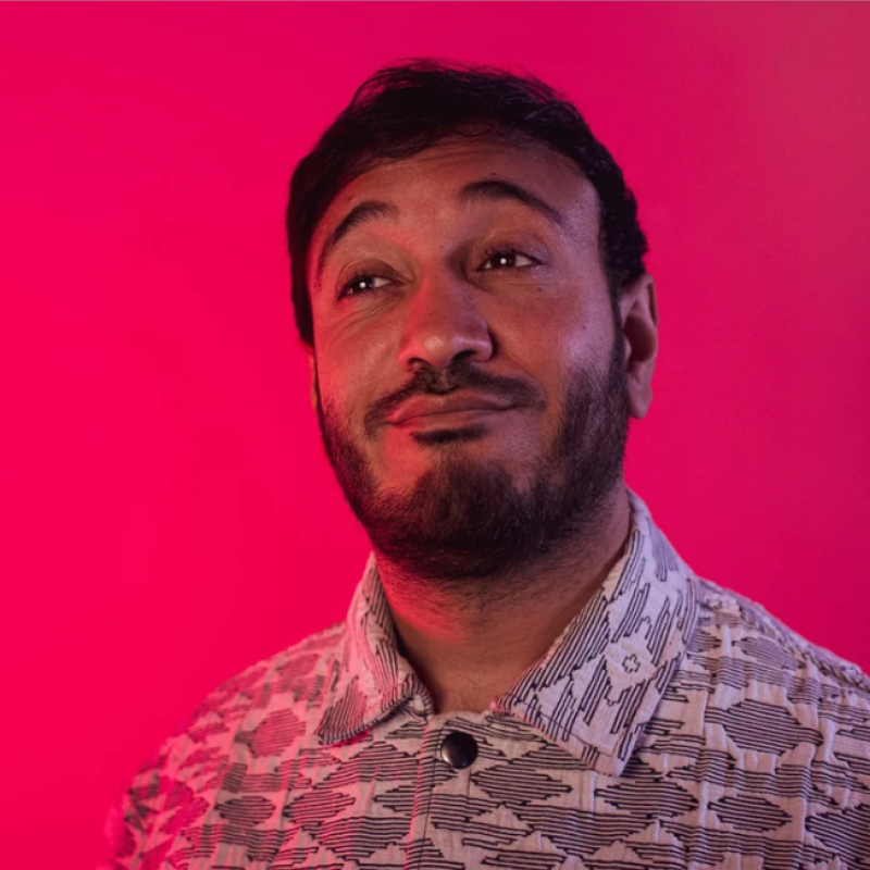 Bilal stands in front of a pink background, in a geometric shirt, looks with a wry smile into the distance