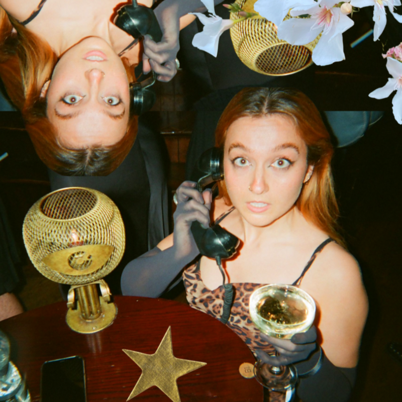 Em holding a black landline phone wearing black opera gloves and a leopard print dress. They hold a glass of champagne in their other hand.