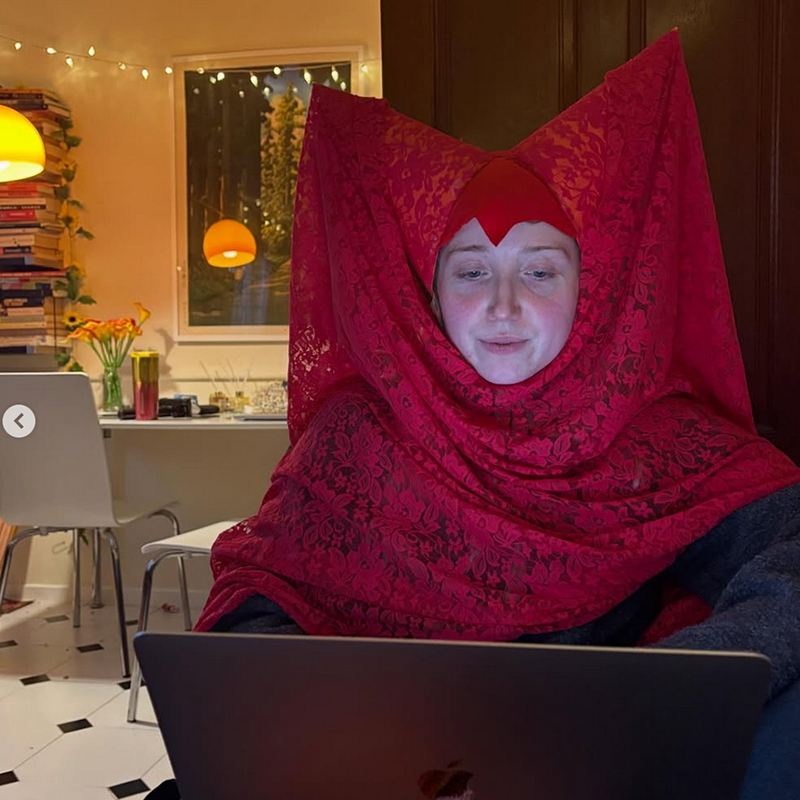 Bebe wearing a medieval inspired red headdress sat at a laptop.