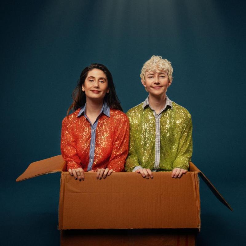 The duo Britney wearing sparkly jackets are sat in a cardboard box under a spotlight.